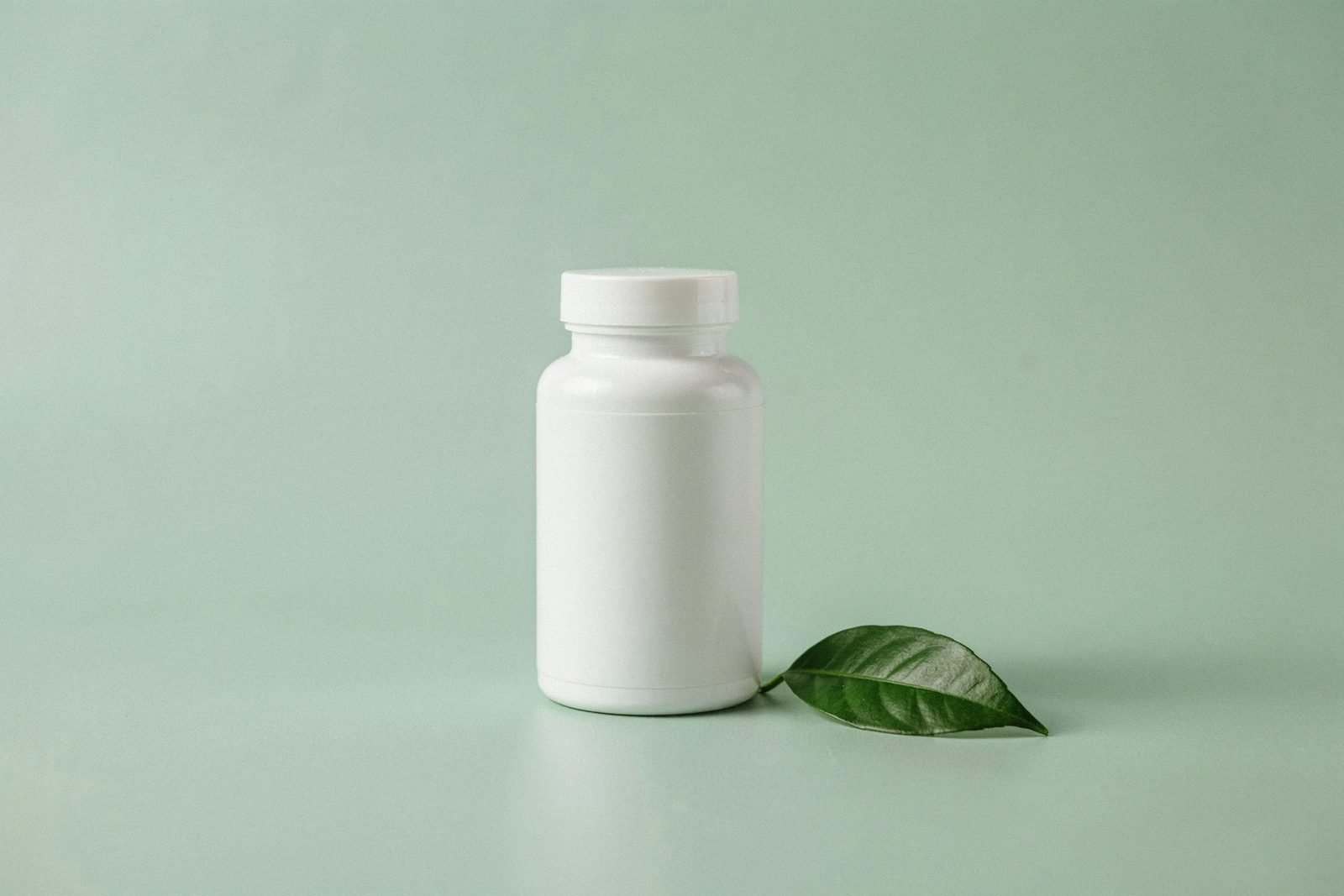Flacon blanc de complément alimentaire posé sur un fond vert d'eau, à côté d'une feuille verte
