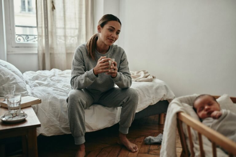 Jeune maman en sweat gris assise au bord de son lit avec une tasse, son nouveau-né endormi dans le berceau à côté