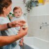 Parent tenant le pot de baume visage Pototam près de bébé, dans une salle de bain Parent tenant le pot de baume visage Pototam près de bébé, dans une salle de bain
