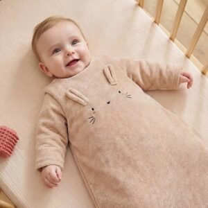 Bébé souriant allongé sur le dos dans un lit à barreaux, en gigoteuse à manches en velours côtelé beige Vertbaudet