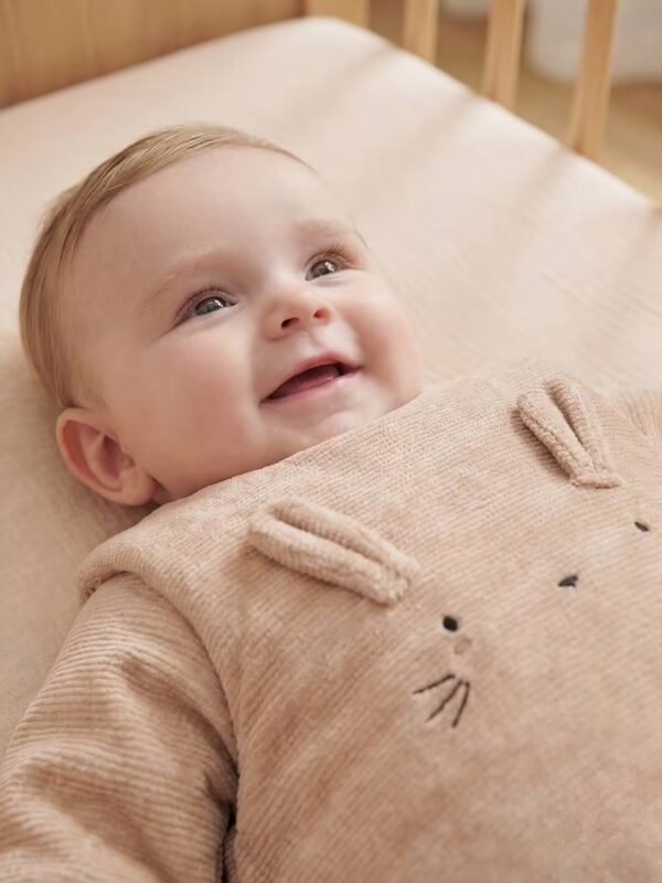 Bébé allongé en gigoteuse velours côtelé Vertbaudet Bébé souriant allongé sur le dos dans son lit, en gigoteuse Vertbaudet en velours côtelé beige à oreilles de lapin