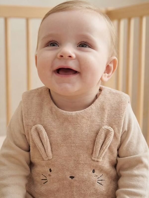 Bébé assis en gigoteuse velours côtelé Vertbaudet Bébé souriant assis dans son lit à barreaux, en gigoteuse Vertbaudet en velours côtelé beige avec oreilles et moustaches de lapin brodées